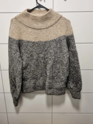 Beige och grå stickad tröja STOCKH LM - Mysig stickad tröja från STOCKH LM i tvåfärgad design med beige upptill och grå nedtill. Tröjan har bred polokrage, ribbade muddar och avslappnad passform. Perfekt för kalla dagar och enkel att styla med jeans eller kjol.