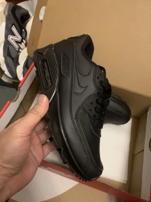Nike Air Max 90 helsvarta sneakers - Snygga helsvarta Nike Air Max 90 sneakers i läder och syntetmaterial. Klassisk siluett med synlig Air-enhet i sulan, svarta skosnören och diskreta detaljer. Perfekta för dig som gillar en clean och tidlös look.