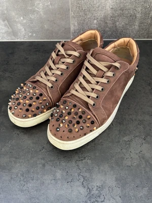 Christian Louboutin  - Snygga och exklusiva sneakers från Christian Louboutin i brun mocka med beige snören och den ikoniska detaljen med dekorativa nitar över tån. Skorna har en klassisk låg sneaker-modell med vita gummisulor och vadderat skaft för en bekväm passform. 🤎✨ Hör av er vid minsta fundering 💭 