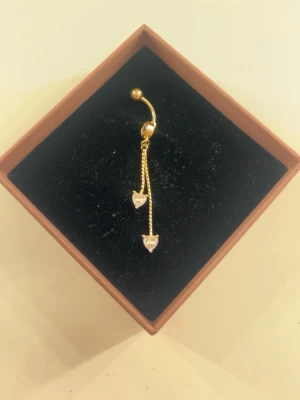 Guldfärgad navelpiercing med hjärtan - Snygg guldfärgad navelpiercing med två tunna kedjor och gnistrande hjärtformade hängen i klar sten. Köpt utomlands. Rengör smycket innan jag postar!💓💓