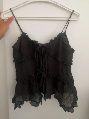 Svart volangig spaghettitopp - Svart topp med volangiga kanter och tunna axelband. Toppen har dekorativa knytningar framtill och ett lätt transparent, vågigt tyg som ger en edgy och trendig look. Perfekt för att styla lager på lager eller till jeans.