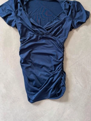 Mörkblå omlotttröja med kort ärm - Snygg mörkblå blus/top i omlottmodell med korta ärmar och lätt rynkade sidor. Tröjan har en djup v-ringning och är tillverkad i ett mjukt, glansigt material som ger en elegant känsla. Perfekt för dig som vill ha en stilren och trendig look. (lång i modellen)