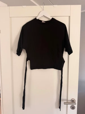Svart croppad topp med knytband Nelly - Säljer en svart croppad topp med korta ärmar och långa knytband i midjan. Perfekt att styla med höga jeans eller kjol för en trendig look. T-shirten har en enkel design och är gjord i mjukt material.