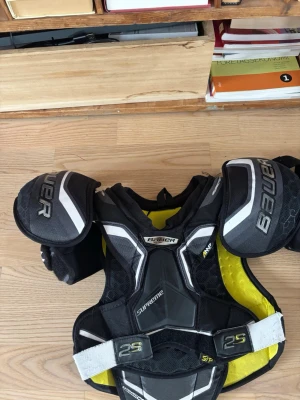 Bauer Supreme axelskydd svart/gul - Säljer ett par Bauer Supreme axelskydd i svart och gul med vita detaljer. Skydden har vadderade axelpartier och bröstskydd, justerbara band och meshpaneler för ventilation. Perfekt för hockey eller annan kontaktsport.