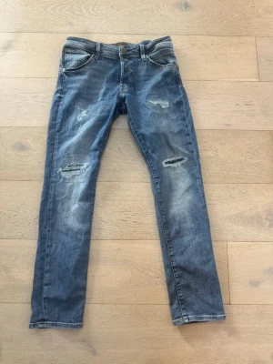 Blå slitna slim fit jeans - Säljer ett par blå slim fit jeans med slitningar och hål på benen för en trendig look. Jeansen har fem fickor, knappgylf och är tillverkade i ett klassiskt denimtyg. Perfekta för dig som gillar en avslappnad och cool stil.