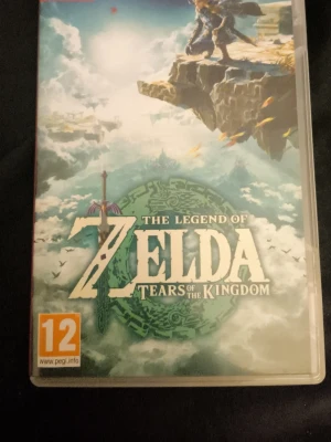The Legend of Zelda: Tears of the Kingdom - Nintendo Switch-spelet The Legend of Zelda: Tears of the Kingdom. Omslaget har en episk illustration med Link på en klippa och svärdet i förgrunden. Färgerna är mestadels blå, grön och vit med fantasy-vibbar. PEGI 12.