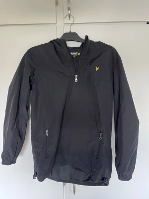 Svart vindjacka med huva från Lyle & Scott - Snygg svart vindjacka från Lyle & Scott med halv dragkedja framtill, huva och två fickor med dragkedja. Jackan har det klassiska gula logomärket på bröstet och elastiska muddar vid ärmsluten. Perfekt för blåsiga dagar.