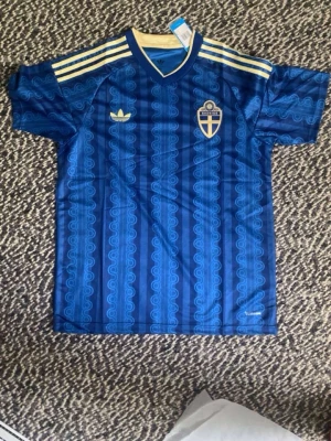Blå Sverige Adidas matchtröja - Säljer en blå matchtröja från Adidas med Sveriges landslagsemblem på bröstet och klassiska gula ränder på axlarna. Tröjan har ett mönstrat tyg, korta ärmar och rund hals. Perfekt för dig som vill stötta Sverige på match eller träning.