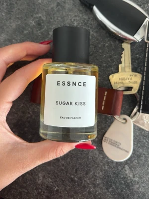 ESSNCE Sugar Kiss Eau de Parfum - ESSNCE Sugar Kiss Eau de Parfum är en stilren parfym i en rund glasflaska med svart matt kork. Doften har en söt och fräsch vibe, perfekt för dig som gillar moderna och trendiga parfymer. Flaskan har en enkel vit etikett med svart text. Aldrig använd, helt ny. 