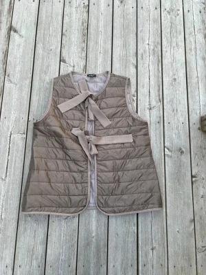 Beige quiltad dunväst med knytband - Stilren beige dunväst med quiltat mönster och två breda knytband framtill. Västen är ärmlös, har rund halsringning och raka kanter. Perfekt för lager-på-lager och ger en avslappnad men trendig look.