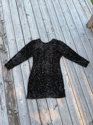 Svart glittrig paljettklänning - Säljer en svart kortklänning täckt av glittriga paljetter. Klänningen har lång ärm och v-ringning i ryggen, vilket ger en snygg och festlig look. Perfekt för dig som vill sticka ut och glänsa på nästa event!