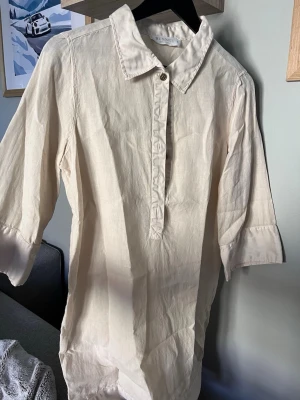 Beige linneskjorta med trekvartsärm - Stilren beige blus i linnematerial med klassisk krage och knappslå framtill från busnel. Blusen har trekvartsärmar med breda manschetter och en avslappnad passform som ger en chill vibe. Perfekt att styla med jeans eller kjol för en enkel men snygg look.