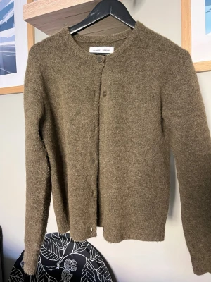 Olivgrön stickad kofta - Mysig olivgrön stickad kofta med rund halsringning och knäppning framtill. Koftan har långa ärmar och är tillverkad i ett mjukt, fluffigt material som passar perfekt för kyligare dagar. Enkel och stilren design som är lätt att matcha.