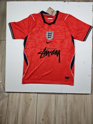 Röd England fotbollströja Stüssy x Nike - Snygg röd England fotbollströja i samarbete mellan Stüssy och Nike. Tröjan har korta ärmar, svart Stüssy-logga framtill, broderat England-emblem och svarta detaljer längs sidorna och ärmsluten. Tillverkad i lätt och ventilerande material, perfekt för träning eller match.