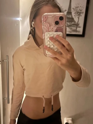 Råsa cropped hoodie med huva - Säljer en beige cropped hoodie med huva och dragsko i midjan. Hoodien har långa ärmar och en enkel, stilren design utan synligt tryck eller logga. Perfekt för en avslappnad och trendig look.