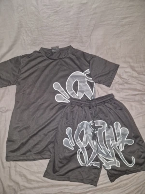 Svart t-shirt med graffiti-tryck - Cool svart t-shirt med stort vitt graffiti-inspirerat tryck på sidan. T-shirten har rund hals och korta ärmar, perfekt för en avslappnad streetwear-look. Materialet känns mjukt och lätt, troligen polyester eller bomull.