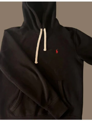 Svart hoodie från Polo Ralph Lauren - Säljer en stilren svart hoodie från Polo Ralph Lauren med klassisk logga på bröstet. Den har en justerbar huva med vita snören och en praktisk magficka. Perfekt för en avslappnad look. Höjd: 59cm och bredd: 48cm