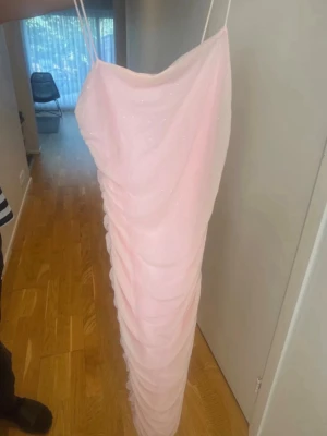 Ljusrosa långklänning med smala band - Elegant ljusrosa långklänning med smala axelband och draperad design. Klänningen har en glittrig yta och är figurnära med en mjuk, romantisk look. Perfekt för speciella tillfällen och sommarens fester.