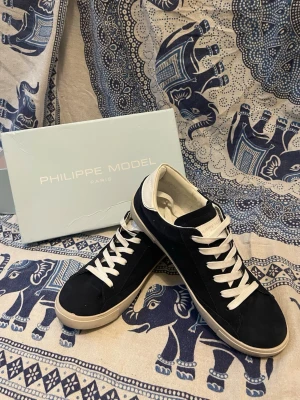 Philippe Model blåa sneakers - Snygga blåa sneakers från Philippe Model med vita detaljer och vita skosnören. Skorna har en klassisk låg siluett och är tillverkade i mocka och läder. Perfekta för dig som vill ha en stilren och trendig look.