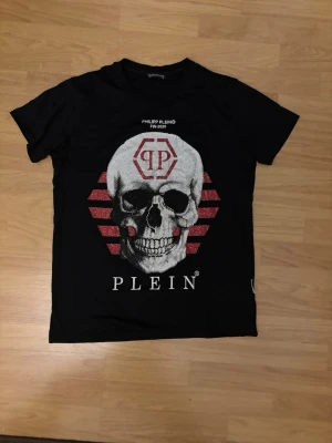 Svart t-shirt med dödskalle från Plein - Cool svart t-shirt från Philipp Plein med stor dödskalle framtill och röda detaljer. T-shirten har korta ärmar, rund hals och tryck med Plein-logga. Perfekt för dig som gillar edgy streetwear och vill sticka ut.