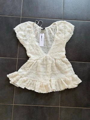 Krämvit spetsblus från Gina Tricot - Supersöt krämvit blus från Gina Tricot i babydoll-modell med spetsdetaljer och volangkant nedtill. Toppen har v-ringning och korta ärmar, perfekt för en romantisk och trendig look. Passar dig som gillar feminina detaljer och lätt material.