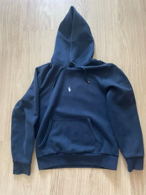 Marinblå hoodie från Polo Ralph Lauren - Snygg marinblå hoodie från Polo Ralph Lauren med klassisk huva och dragsko. Har en broderad vit logga på bröstet och en stor känguruficka framtill. Tillverkad i mjukt bomullsmaterial som är skönt att bära. Perfekt för en avslappnad och stilren look.