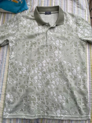 Grönvit polotopp från Dior - Säljer en snygg polotopp från Dior i ljusgrönt och vitt med ett diskret mönster och klassisk krage. Toppen har knappar framtill och är perfekt för dig som gillar exklusiv och trendig stil. Materialet känns mjukt och andas bra.