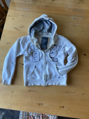 Vit hoodie Abercrombie & Fitch - Vit zip-hoodie från Abercrombie & Fitch med ikoniska FITCH 92-applikationer på bröstet. Modellen är lite lätt sliten/vintage look som är väldigt vanligt vid A&F äldre modeller.  Skick: bra skick lite lätta skavanker som tex att löparen på dragkedjan är lite trasig men dragkedjan funkar som vanligt bara utan en grej att hålla i. Storleken är i M men passar lika bra på S och passformen är normal. Hör gärna av er vid funderingar. Pris ej hugget i sten. 