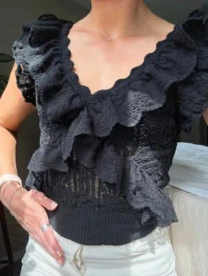 Svart spetsblus med volanger - Supersnygg svart blus i spets med breda volanger framtill och längs axlarna. Djup v-ringning och ribbad midja ger en feminin och trendig look. Perfekt att styla med både jeans och kjol för en cool outfit.