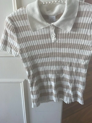 Randig ribbad topp från Lindex - Säljer en ribbad topp från Lindex i beige och vitt med horisontella ränder. Toppen har korta ärmar, klassisk krage och är tillverkad i mjuk bomull. Perfekt för dig som gillar en enkel men snygg stil.