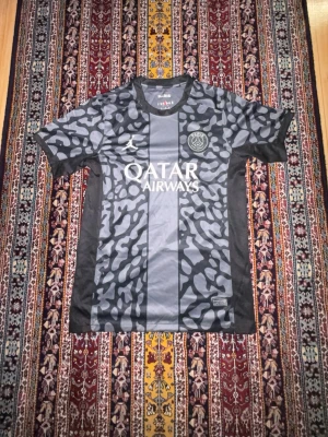 Svart PSG x Jordan matchtröja - Snygg svart och grå PSG matchtröja från Jordan med camo-liknande mönster och vita detaljer. Tröjan har Paris Saint-Germain-logga på bröstet och Qatar Airways tryckt på magen. Tillverkad i lätt och ventilerande material, perfekt för fotboll eller träning.