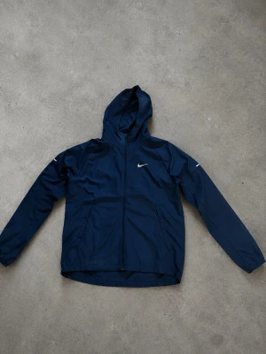 Mörkblå Nike vindjacka med huva - Snygg mörkblå vindjacka från Nike med huva och dragkedja framtill. Jackan har två sidofickor och den klassiska Nike-loggan på bröstet. Tillverkad i lätt och vindtätt material, perfekt för blåsiga dagar.