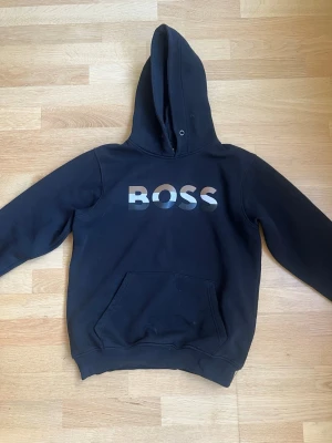 Svart hoodie med BOSS-tryck - Snygg svart hoodie med stor huva och magficka framtill. På bröstet står det BOSS i ett metallicfärgat tryck. Hoodien har ribbade muddar vid ärmslut och nederkant. Perfekt för en chill och avslappnad stil.