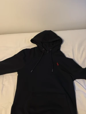 Svart hoodie från Polo Ralph Lauren - Snygg svart hoodie från Polo Ralph Lauren med klassisk röd logga broderad på bröstet. Hoodien har dragsko i huvan och en stor magficka framtill. Tillverkad i mjukt bomullsmaterial som är perfekt för chill dagar.