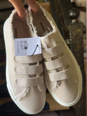 Beige sneakers med kardborreband - Snygga beige sneakers med tre breda kardborreband och råa kanter för en trendig look. Skorna har en vit sula och är tillverkade i textilmaterial. Perfekta för en avslappnad och stilren outfit.