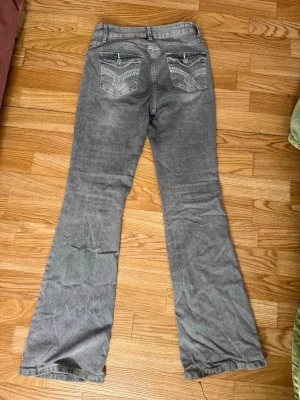 Grå bootcut jeans med detaljer - Säljer ett par grå bootcut jeans med snygga broderade detaljer och knappar på bakfickorna. Jeansen har hög midja och utsvängda ben, vilket ger en retro vibe. Perfekta för dig som vill sticka ut lite extra med coola detaljer.