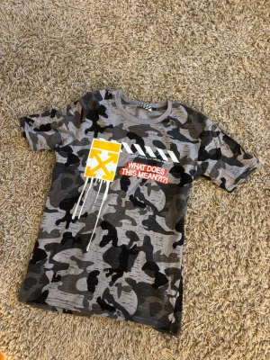 Camouflage t-shirt från Off-White - Snygg t-shirt från Off-White med grått och svart camouflage-mönster. Tryck på bröstet med texten 'WHAT DOES THIS MEAN?!?' och Off-White-logga i gult och vitt. Rund hals och korta ärmar. Perfekt för dig som gillar streetwear och statement-plagg.