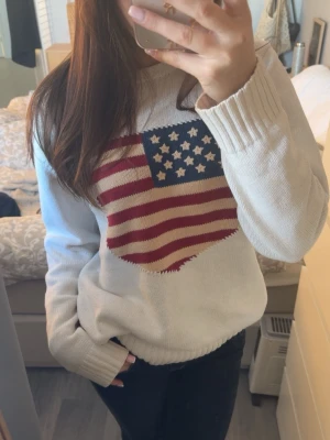 Vit stickad tröja Gant - Vit stickad tröja med stor USA-flagga i rött, blått och vitt på bröstet. Tröjan har rund halsringning och ribbade muddar vid ärmslut och nederkant. Perfekt för dig som gillar en avslappnad och trendig stil.
