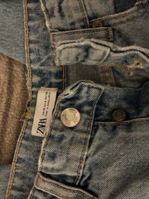 Ljusblå raka jeans från ZARA - Säljer ett par ljusblå jeans från ZARA med rak passform och klassisk femficksdesign. Jeansen har slitna detaljer vid bensluten och är tillverkade i denim. Perfekta för en avslappnad och trendig look.