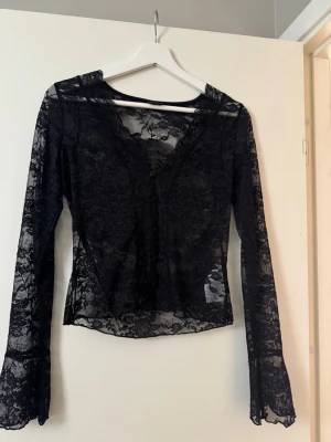 Svart spetsblus med v-ringning - Snygg svart långärmad blus i genomskinlig spets med v-ringad hals. Blusen har en figurnära passform och är perfekt för att skapa en trendig lager-på-lager-look. Tunna spetsdetaljer över hela toppen ger en elegant vibe.
