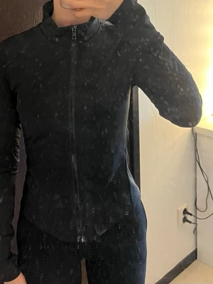 Svart sportjacka med dragkedja - Säljer en svart sportjacka med hög krage och hel dragkedja framtill. Jackan är figursydd och har långa ärmar, perfekt för träning eller löpning. Materialet är stretchigt och följsamt för maximal rörelsefrihet.