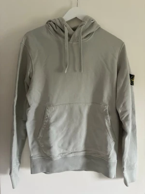 Stone Island Hoodie - Snygg ljusgrå hoodie från Stone Island med klassisk patch på vänster ärm. Hoodien har huva med dragsko och en stor magficka framtill. Tillverkad i mjukt bomullsmaterial som är skönt att bära. Perfekt för en avslappnad streetwear-look. Storlek S