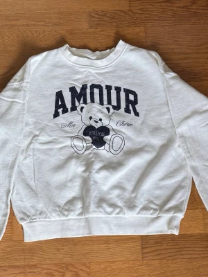 Vit sweatshirt med björntryck Kappahl - Vit sweatshirt från Kappahl med gulligt björntryck med texten ”Amour” framtill. Mjuk, bekväm och tunn som är perfekt för sommaren, med ribbade muddar. STORLEK: 146/152