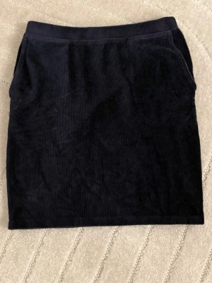 Svart manchesterkjol med fickor - Säljer en svart kortkjol i manchester med fickor på sidorna. Kjolen har en enkel och stilren design som passar perfekt till olika outfits. Materialet är mjukt och ger en snygg struktur.