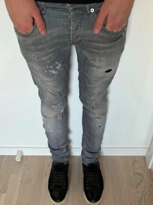 Grå dondup jeans  - PASSAR 33 32!!!Säljer ett par grå skinny jeans med slitningar och hål på benen. Jeansen har en tvättad look med färgstänk och trasiga detaljer för en trendig streetstil. Femficksmodell och låg midja. Perfekta för dig som gillar en avslappnad och edgy vibe.