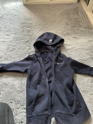 Marinblå hoodie från Sail Racing - Snygg marinblå hoodie från Sail Racing med dragkedja framtill och två stora fickor. Hoodien har logga på bröstet och på huvan, samt mjukt material på insidan. Perfekt för en avslappnad och sportig stil.