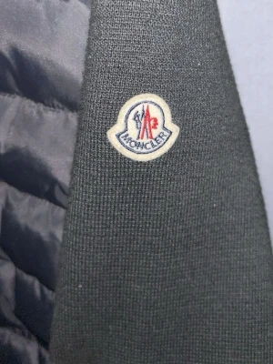 Svart pufferjacka från Moncler - Snygg svart pufferjacka från Moncler med ribbad krage och Moncler-logga på ärmen. Jackan har dragkedja framtill och quiltade partier som ger en modern look. Perfekt för dig som vill ha både stil och värme under kyliga dagar. Stolek S/M
