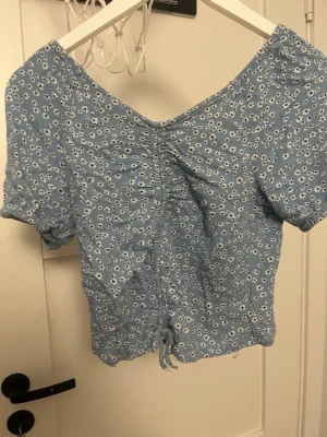 Blå blommig omlottopp med snörning - Supersöt blå omlottopp med små vita blommor och snörning framtill. Toppen har korta puffärmar och v-ringad. Materialet känns lätt och luftigt.