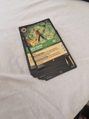 Disney lorcana emerald 47st - En hög med samlarkort från ett spel, med färgstarka illustrationer och text på engelska. Korten är rektangulära och gjorda av papper eller kartong. Perfekt för dig som gillar strategispel och samlarkort.