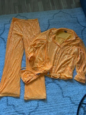 Glittrigt orange set med paljetter - Säljer ett riktigt snyggt set i orange med massor av paljetter. Setet består av långärmad topp med knappar, matchande byxor med utsvängda ben och en liten topp. Materialet är stretchigt och skimrar i ljuset – perfekt för dig som vill sticka ut.